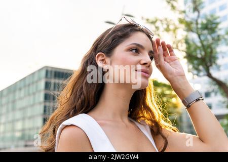 Low angle of self assured young female with long brown hair adjusting sunglasses and looking away against modern glass skyscrapers Stock Photo