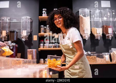 Glad young African American woman in striped apron and glasses looking at camera with smile and putting glass box of dried mango on shelf during work Stock Photo