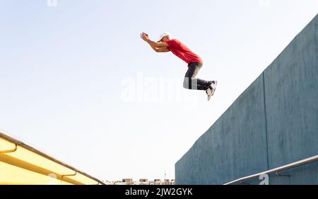 Active male hipster jumping from edge of grey building while doing parkour trick in sunny day Stock Photo