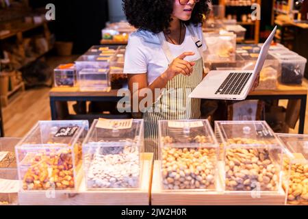 Cropped unrecognizable young African American woman in uniform and glasses with curly hair checking goods in glass boxes and using laptop while workin Stock Photo