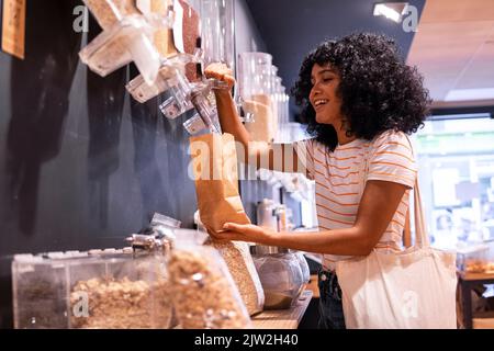 Cheerful young African American woman in casual clothes with curly hair smiling and using dispenser to fill paper bag while visiting eco friendly groc Stock Photo
