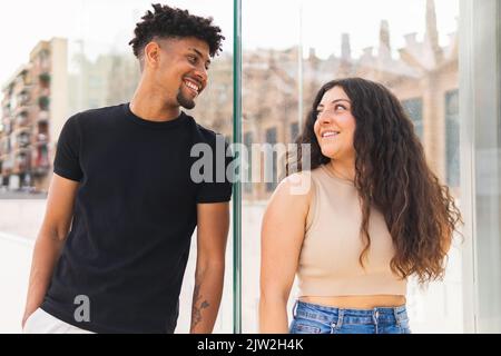Smiling young couple looking at each other while standing by glass wall in city Stock Photo