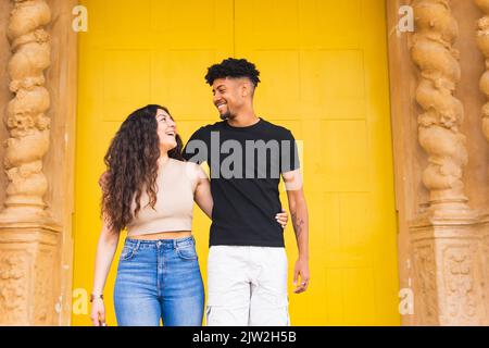 Smiling young couple looking at each other while standing with arm around against yellow door Stock Photo