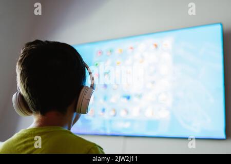 Back view of anonymous boy in wireless headphones playing interesting video game on TV in light living room Stock Photo