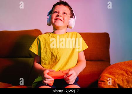 Delighted boy in wireless headphones with gamepad while playing video game on couch in living room Stock Photo