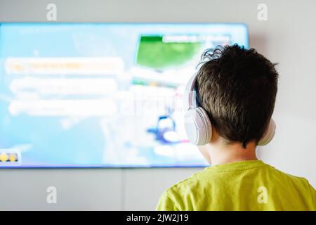 Back view of anonymous boy in wireless headphones playing interesting video game on TV in light living room Stock Photo