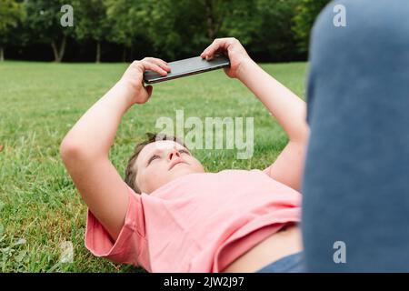Kid lying down on grassy lawn and playing videogame on mobile phone on weekend day in park Stock Photo