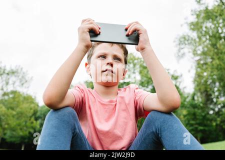 Child in pink t shirt and leggings sitting on grassy lawn and watching video on cellphone on weekend day in park Stock Photo