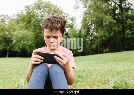 Upset child in pink t shirt and leggings sitting on grassy lawn and watching video on cellphone on weekend day in park Stock Photo