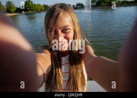 Teenage girl looking at camera while taking self portrait on smartphone on paved waterfront near rippling river on summer day Stock Photo