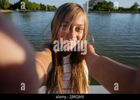 Teenage girl looking at camera while taking self portrait on smartphone on paved waterfront near rippling river on summer day Stock Photo