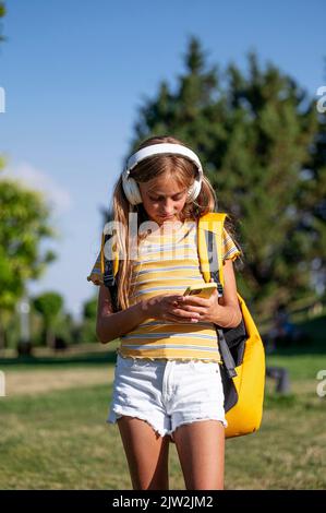 Focused teenage girl with backpack listening to music in wireless headphones while surfing cellphone in park on sunny summer day Stock Photo