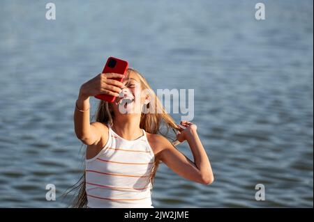 Cheerful teenage girl taking self portrait on smartphone on paved waterfront near rippling river on summer day Stock Photo