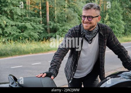 Man biker middle aged motorcyclist smiling stands with a motorcycle on the side of the road in the forest Stock Photo
