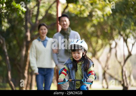 little asian girl riding bike in city park with parents in background Stock Photo
