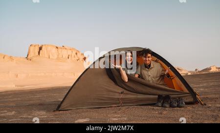 Happy couple sitting in tent during trip in Lut desert with rough rocky cliffs on sunny summer day in Iran Stock Photo