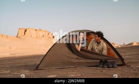 Happy couple sitting in tent during trip in Lut desert with rough rocky cliffs on sunny summer day in Iran Stock Photo