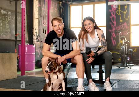 Happy young man and woman with Bull Terrier dog smiling and looking at camera while sitting on bench during break in gym Stock Photo