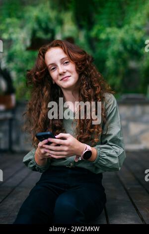 Positive young female with long red curly hair and freckles in casual clothes smiling and looking at camera while messaging on mobile phone sitting on Stock Photo