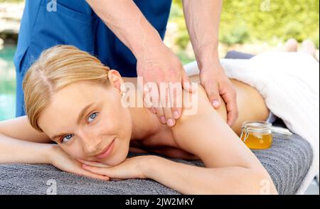 Happy blond female client smiling and looking at camera while crop masseur doing massage during spa session on resort Stock Photo