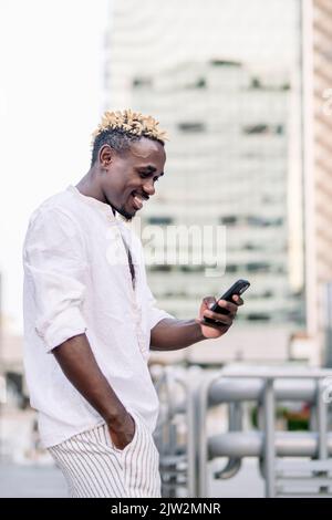 Side view of glad black male in t shirt looking at screen with smile while texting on cellphone in daytime on city street Stock Photo