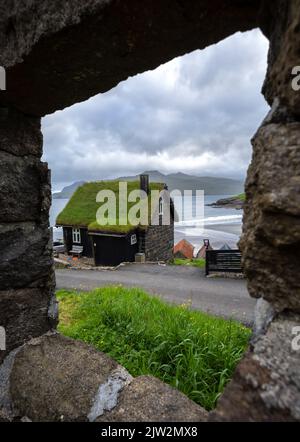 Picturesque view of stone house with mossy roof located against waving sea and cloudy sky behind window of old building on stormy day on Faroe Islands Stock Photo