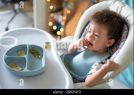 Baby boy eating by himself in his high chair at home. Adorable one year old baby having a meal holding food in his hands and eating at home Stock Photo