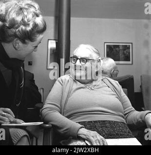 1965, historical, an elderly lady sitting in a chair in the lounge area of an old peoples home, being interviewed by a female journalist, perhaps telling her lifestory. Stock Photo