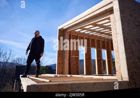 Male builder building wooden frame house. Bald man standing on construction site, inspecting quality of work on sunny day with blue sky on background. Stock Photo