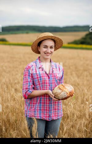 A female farmer in a straw hat and checkered shirt holds fragrant bread in her hands on a ripe wheat field. The smell of freshly baked bread. Stock Photo