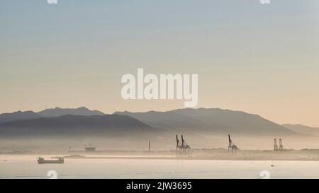 Sunset in the port of Malaga with fog caused by humidity with cranes, ships sailing and mountains behind Stock Photo