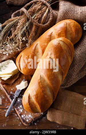 Rustic breakfast table setting. Two freshly baked loaves on wooden cutting board with organic butter and knife. Top view of uncut white bread lying on Stock Photo