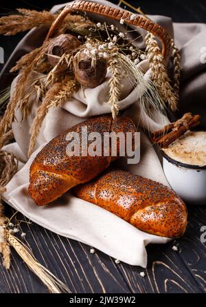 Baked sweet rolls sprinkled with poppy seeds lie on burlap on country table. Two rolls and old enameled cup with coffee and cinnamon near wicker baske Stock Photo
