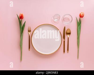 Festive pink table setting. Pink plate with gold cutlery and red tulip on pink background. Aesthetic stylish dinner table setting for Women's day, Mother's Day,wedding or anniversary.Copyspace topview Stock Photo