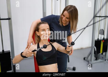 Rehabilitologist physiotherapist assisting woman working on pull cable exercising machine Stock Photo