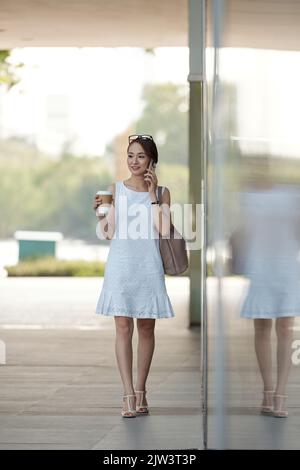 Beautiful young woman talking on phone and drinking coffee when going to work Stock Photo