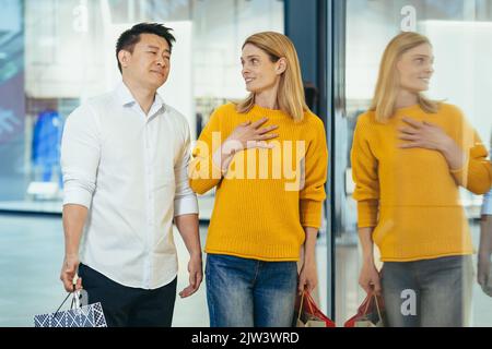 multiracial couple asian man and caucasian woman walking together in mall after shopping with colored bags in hands. walking past shop windows with clothes cheerful family hugging smile. Stock Photo
