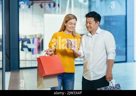 Diverse couple Asian man and blonde woman in clothes supermarket, looking at mobile phone screen, choosing shopping, holding colorful shopping bags and gifts. Stock Photo