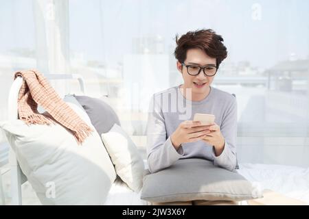 Cheerful young Asian man in glasses reading message in his smartphone Stock Photo