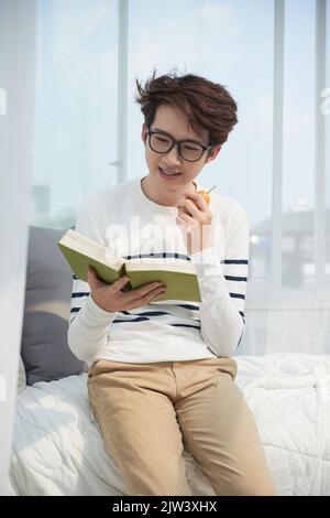 Young smiling Vietnamese man eating a pear and reading an interesting book Stock Photo