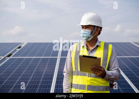 A young Asian engineer holding a tablet stands inspecting an alternative energy solar panel installed in a rural area. Stock Photo