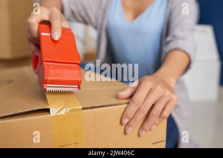 Close-up view of unrecognizable woman sealing cardboard box with adhesive tape while preparing for moving out Stock Photo