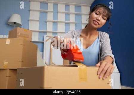 Low angle view of young attractive Asian woman packing carton box with adhesive tape while moving out of apartment Stock Photo
