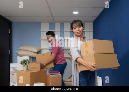 Young Asian woman holding carton boxes in new home and smiling at camera while her husband helping to unpack Stock Photo