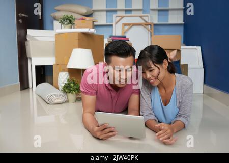 Young Asian couple using digital tablet and smiling happily while lying on the floor in new flat with packed household stuff Stock Photo
