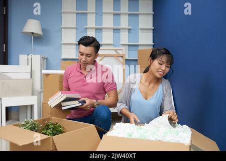 Young happy Asian couple unpacking cardboard boxes with books and household stuff after moving in new apartment Stock Photo