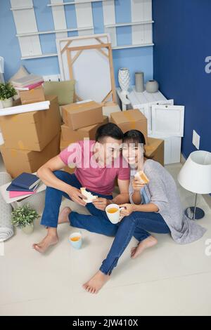 Young Asian couple eating sandwiches and smiling happily while sitting on the floor in messy room with unpacked carton boxes Stock Photo