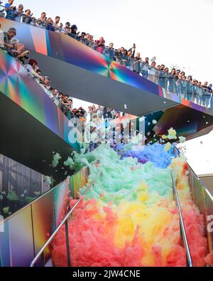 London, UK. 03rd Aug, 2022. People interact with 'Island of Foam: Version XVIII', a colourful, constantly changing foam installation. The installation is a UK premiere from German artist Stephanie Lüning (who is operating the 'foam cannon' at the top of the stairs), transforming Greenwich Peninsula with mountains of rainbow coloured foam. Credit: Imageplotter/Alamy Live News Stock Photo