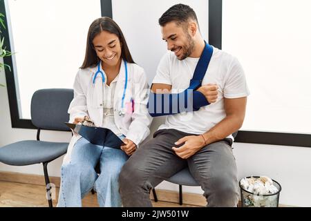 Youg latin injuried man wearing sling talking with the doctor at clinic waiting room. Stock Photo