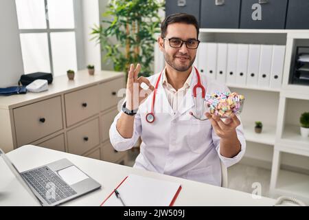 Young hispanic doctor man with beard holding candy doing ok sign with fingers, smiling friendly gesturing excellent symbol Stock Photo
